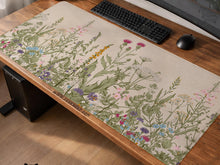 A desk mat with a botanical meadow design featuring wildflowers, leafy stems, and soft neutral colors.