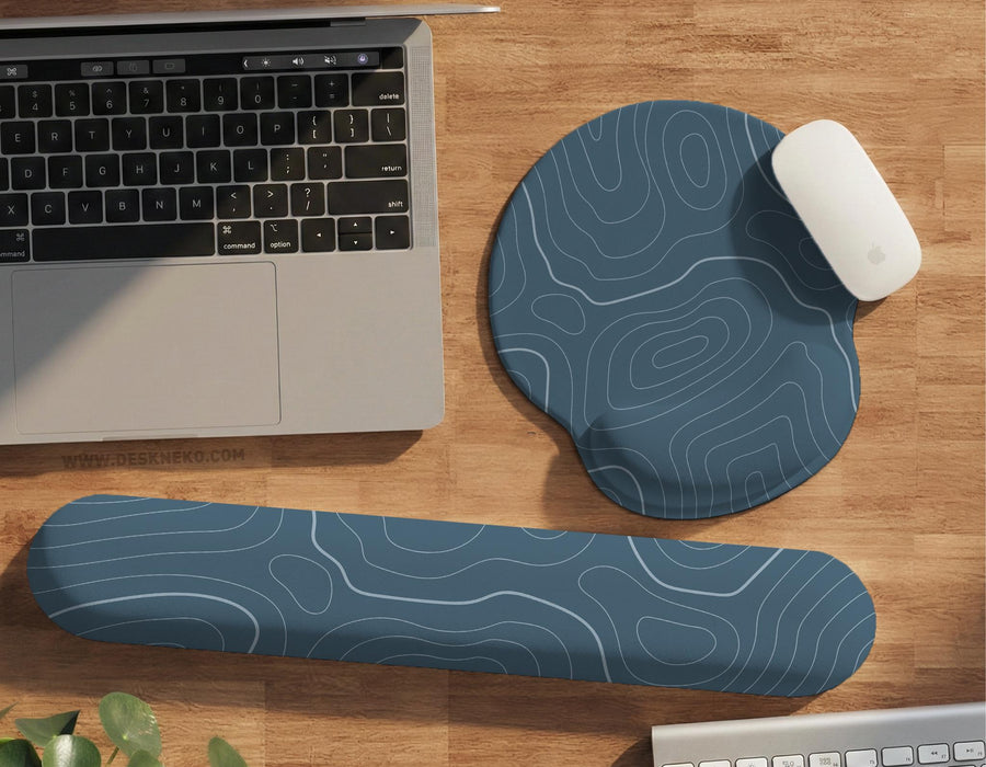 Muted blue desk mat with thin topographic line art pattern shown with matching mousepad and keyboard wrist rest options.