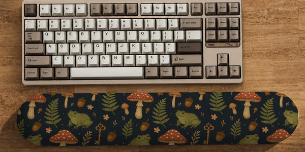 Dark botanical desk mat options with frogs, mushrooms, ferns, and acorns in brown and green tones, plus a matching keyboard wrist rest.