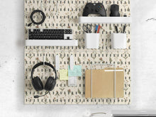 Cream pegboard wall organizer with small floral print, sage green stems, black slot layout, shelves, hooks, clips, and containers.