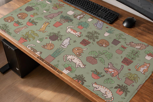 A desk mat with a repeating pattern of cats, leafy houseplants, cactus pots, and small plant-shop style details in sage green.