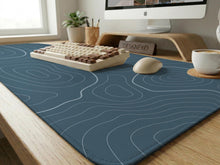 A slate blue desk mat with thin contour line map art on a light background.