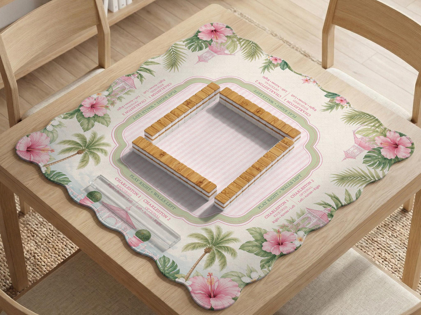 Pink mahjong mat with a scalloped edge design, palm leaves, hibiscus flowers, soft sage accents, and a striped center play area with printed rules around the border.