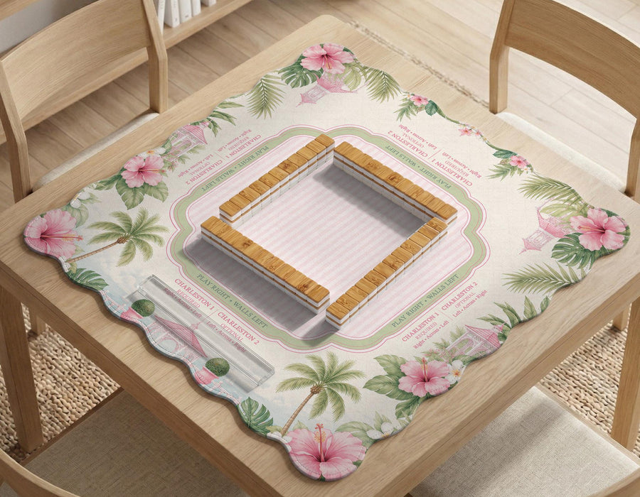 Pink mahjong mat with a scalloped edge design, palm leaves, hibiscus flowers, soft sage accents, and a striped center play area with printed rules around the border.