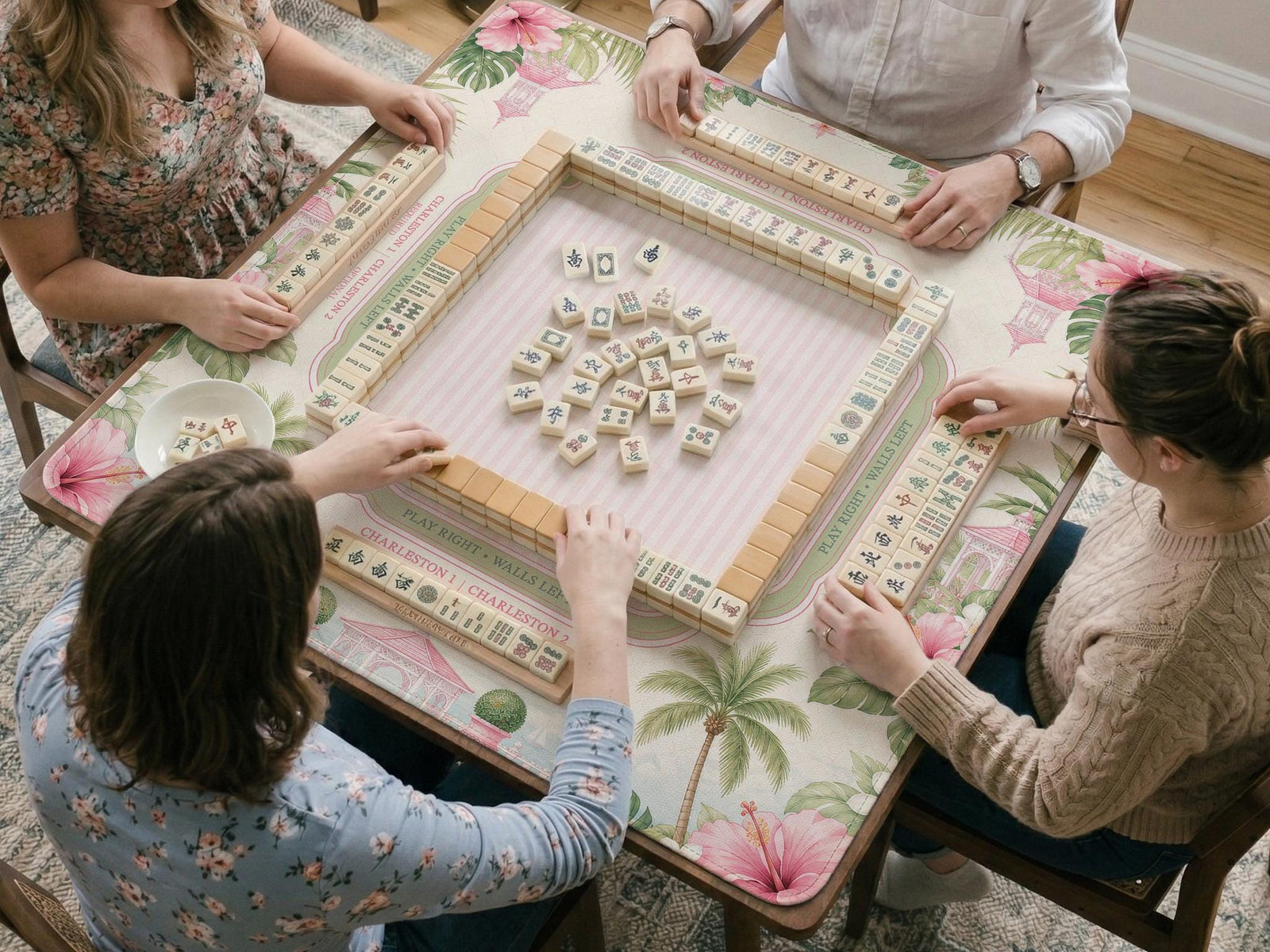 Pink mahjong mat with hibiscus flowers, palm leaves, sage green borders, a cream textured background, and a striped center area for tile play.