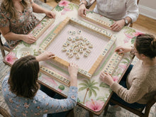 Pink mahjong mat with hibiscus flowers, palm leaves, sage green borders, a cream textured background, and a striped center area for tile play.