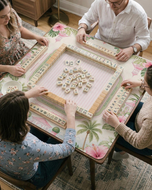 Pink mahjong mat with hibiscus flowers, palm leaves, sage green borders, a cream textured background, and a striped center area for tile play.