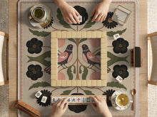 Beige mahjong mat with a folk floral design, a bird, blush pink fruit, olive botanical leaves, black flowers, and a soft woven-look background.