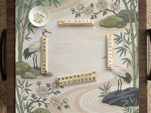 Square mahjong mat with a cream background, sage green cranes, bamboo leaves, peony branches, stones, and thin linework.