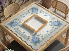 A square blue mahjong mat with hydrangea flowers, cream blossoms, sage leaves, and vines around a plain center play area. The edge is scalloped.