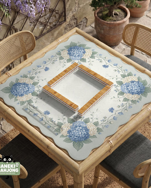 A square blue mahjong mat with hydrangea flowers, cream blossoms, sage leaves, and vines around a plain center play area. The edge is scalloped.