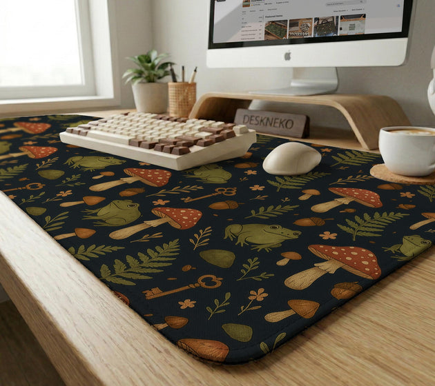 Dark forest-themed desk mat with an all-over pattern of frogs, mushrooms, fern leaves, acorns, tiny flowers, and antique keys.