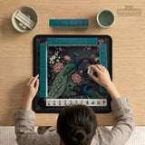 Square black mahjong mat with a gold peacock, blush flowers, and dark botanical artwork on a dark background.