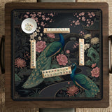 Square black mahjong mat with a gold peacock, blush flowers, and dark botanical artwork on a dark background.