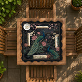 Square black mahjong mat with a gold peacock, blush flowers, and dark botanical artwork on a dark background.