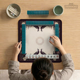 Square mahjong mat with a dark plum border, ivory floral pattern, muted sage leaves, and a decorative peacock garden design.