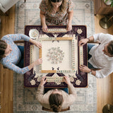 Square mahjong mat with a dark plum border, ivory floral pattern, muted sage leaves, and a decorative peacock garden design.