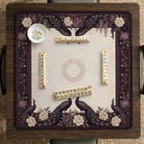 Square mahjong mat with a dark plum border, ivory floral pattern, muted sage leaves, and a decorative peacock garden design.
