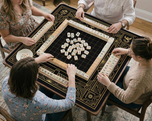 Black and gold square mahjong mat with a scalloped edge, geometric border, thin line details, fan motifs, and dragon and phoenix accents around an open center.
