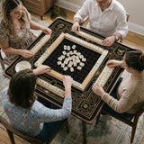 Black and gold square mahjong mat with a scalloped edge, geometric border, thin line details, fan motifs, and dragon and phoenix accents around an open center.