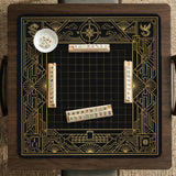 Black and gold square mahjong mat with a scalloped edge, geometric border, thin line details, fan motifs, and dragon and phoenix accents around an open center.