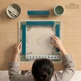 Square mahjong mat with a sage green bamboo and mountain design, cream center, scalloped border details, and gold line accents.