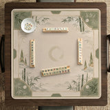 Square mahjong mat with a sage green bamboo and mountain design, cream center, scalloped border details, and gold line accents.
