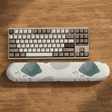 Sage green ergonomic mousepad and keyboard wrist rest with abstract shapes, soft gray details, cream space, topographic contour lines, and leafy branch accents.