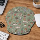 Sage green desk mat with a pattern of lounging cats, leafy plants, cactus pots, and earthy botanical details.