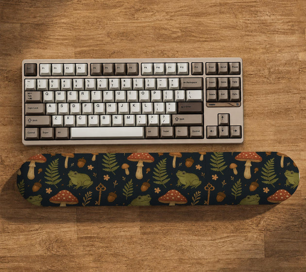 Dark botanical desk mat options with frogs, mushrooms, ferns, and acorns in brown and green tones, plus a matching keyboard wrist rest.