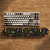 Dark botanical desk mat options with frogs, mushrooms, ferns, and acorns in brown and green tones, plus a matching keyboard wrist rest.