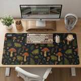Dark forest-themed desk mat with an all-over pattern of frogs, mushrooms, fern leaves, acorns, tiny flowers, and antique keys.