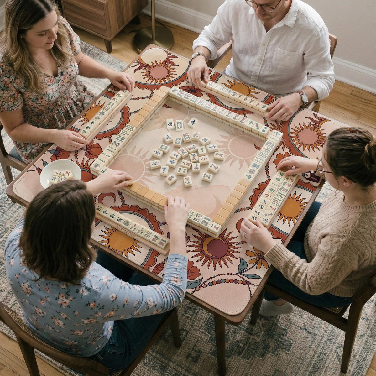 A square mahjong mat with a scalloped edge, sun and moon artwork, terracotta flowers, a soft cream background, and a decorative border around the play area.
