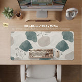 A desk mat with sage green abstract blocks, cream space, gray stone-like shapes, fine topographic contour lines, and small branch details.