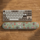 Sage green desk mat with a pattern of lounging cats, leafy plants, cactus pots, and earthy botanical details.
