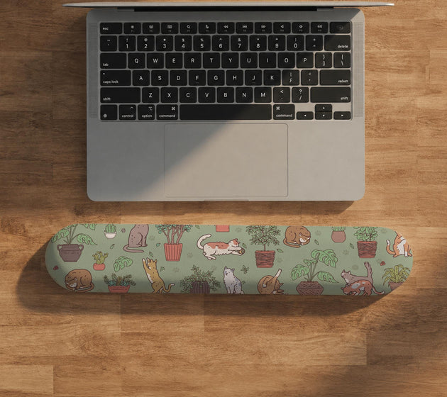 Sage green desk mat with a pattern of lounging cats, leafy plants, cactus pots, and earthy botanical details.