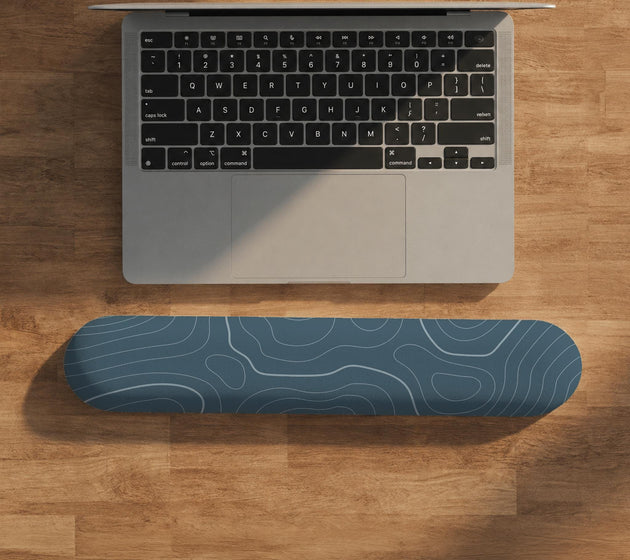 Muted blue desk mat with thin topographic line art pattern shown with matching mousepad and keyboard wrist rest options.
