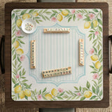 Square yellow mahjong mat with a citrus garden border, green vines, pink blossoms, aqua accents, and a plain center play area.