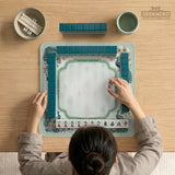Teal square mahjong mat with peacock feathers, soft floral pattern, tassel details, and printed rule guide text in the center.