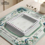 Teal square mahjong mat with peacock feathers, soft floral pattern, tassel details, and printed rule guide text in the center.