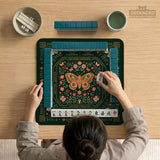 Dark botanical mahjong table mat with a floral border, large moth and butterfly design in the center, gold corner frame, and printed rules layout around the edge.