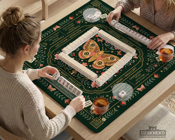 Dark botanical mahjong table mat with a floral border, large moth and butterfly design in the center, gold corner frame, and printed rules layout around the edge.