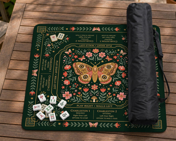 Dark botanical mahjong table mat with a floral border, large moth and butterfly design in the center, gold corner frame, and printed rules layout around the edge.