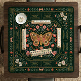 Dark botanical mahjong table mat with a floral border, large moth and butterfly design in the center, gold corner frame, and printed rules layout around the edge.