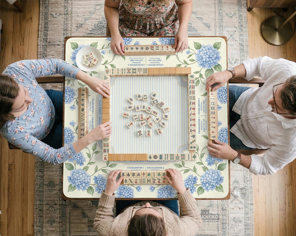 A square mahjong mat with blue hydrangea flowers, green vines, a cream background, a striped center, and printed rules around the border.