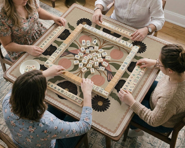 Beige mahjong mat with a botanical floral bird design, blush flowers, olive green leaves, black flower accents, and a warm linen-style background. Scalloped edge option shown.