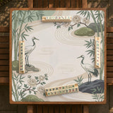 Square mahjong mat with a cream background, sage green cranes, bamboo leaves, peony branches, stones, and thin linework.