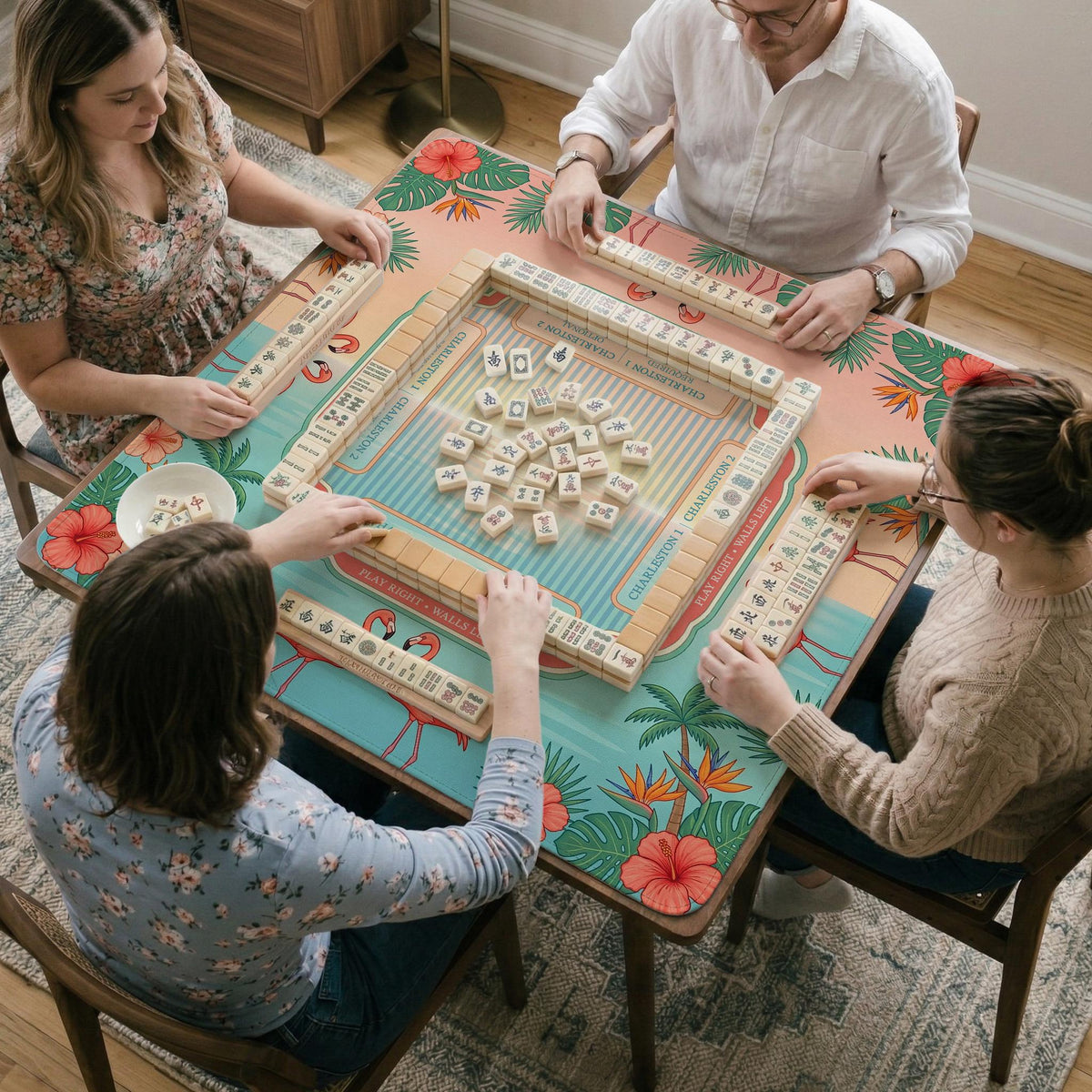 A square mahjong mat with flamingos, hibiscus flowers, palm leaves, and aqua water accents on a coral background with a scalloped edge.