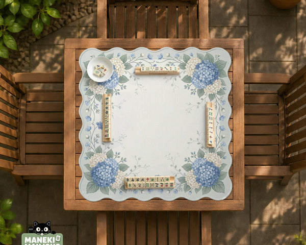 A square blue mahjong mat with hydrangea flowers, cream blossoms, sage leaves, and vines around a plain center play area. The edge is scalloped.