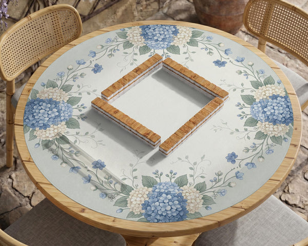 A square blue mahjong mat with hydrangea flowers, cream blossoms, sage leaves, and vines around a plain center play area. The edge is scalloped.